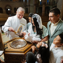 Deacon pours holy water on a baby during baptism as family gathers in a church, with siblings watching closely.