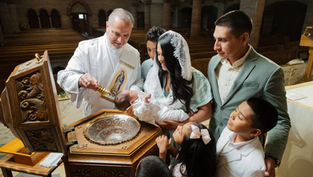 Deacon pours holy water on a baby during baptism as family gathers in a church, with siblings watching closely.