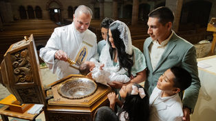 Deacon pours holy water on a baby during baptism as family gathers in a church, with siblings watching closely.