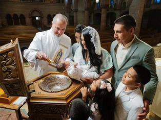 Deacon pours holy water on a baby during baptism as family gathers in a church, with siblings watching closely.