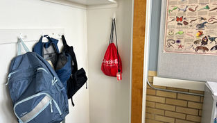 Backpacks are hanging on white hooks in a classroom. A red bag with "Go Red Bag" text is on display. A colorful animal chart is on the wall.