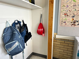 Backpacks are hanging on white hooks in a classroom. A red bag with "Go Red Bag" text is on display. A colorful animal chart is on the wall.