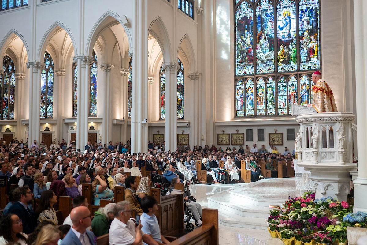 A bishop preaches from the pulpit inside a cathedral filled with a large congregation beneath stained glass windows.