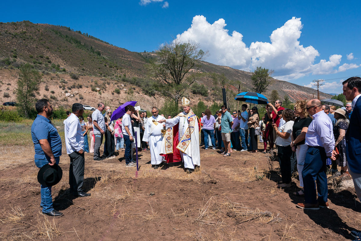 Bishop in ornate vestments blesses a crowd gathered in a dry mountain field, with umbrellas under a bright blue sky.