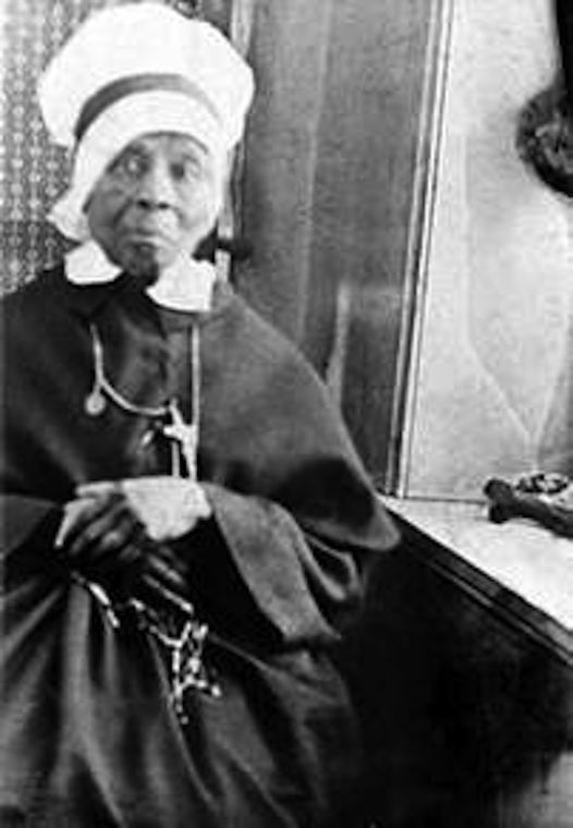 Black-and-white portrait of an elderly woman in a white bonnet and dark dress, seated indoors with a calm expression.