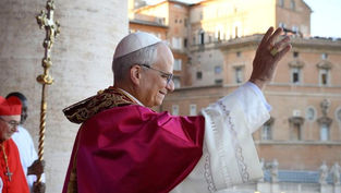 A religious leader in white and red robes waves to a crowd from a veranda. A cardinal stands nearby, with historic buildings in the background.