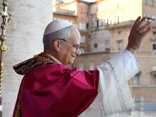 A religious leader in white and red robes waves to a crowd from a veranda. A cardinal stands nearby, with historic buildings in the background.
