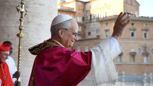 A religious leader in white and red robes waves to a crowd from a veranda. A cardinal stands nearby, with historic buildings in the background.
