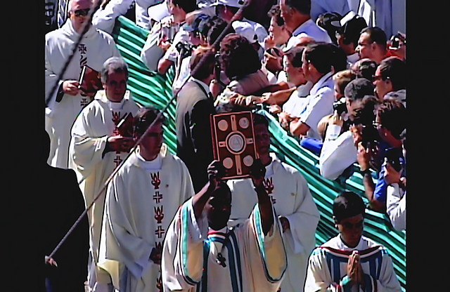 Priests in white robes process with a large book held aloft. A crowd watches from behind green-striped barriers under a bright sky.