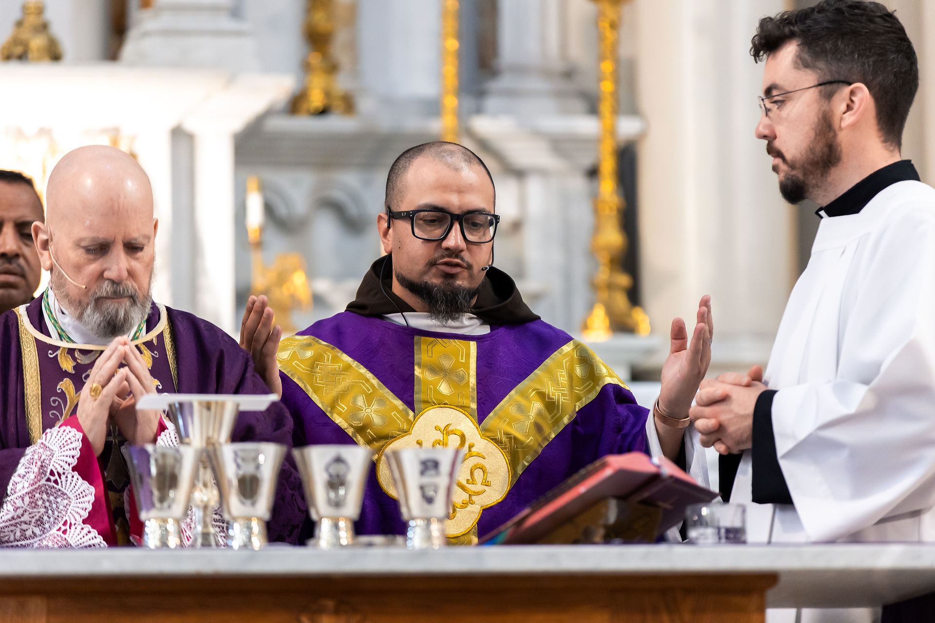 Three clergy in a church, wearing purple and white robes, conduct a ceremony with silver chalices. The setting is ornate and solemn.