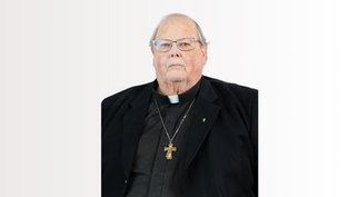 Elderly clergyman in black attire with a cross necklace, neutral expression, light background.