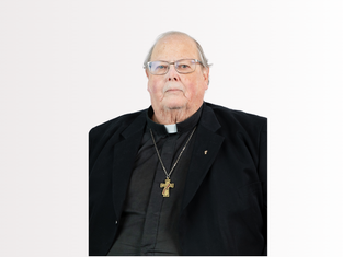 Elderly clergyman in black attire with a cross necklace, neutral expression, light background.