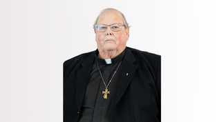 Elderly clergyman in black attire with a cross necklace, neutral expression, light background.