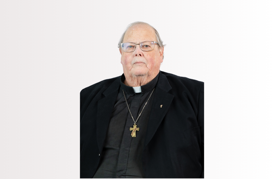Elderly clergyman in black attire with a cross necklace, neutral expression, light background.