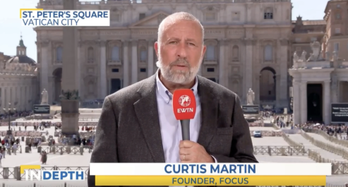 Man holding a red microphone labeled "EWTN" in St. Peter’s Square, Vatican City. Sign reads "Curtis Martin, Founder, FOCUS."