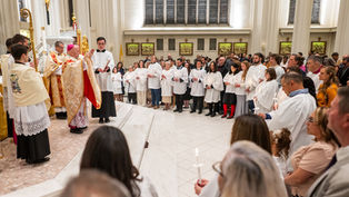 A priest blesses a congregation in white robes holding candles. The ceremony takes place in a bright church with tall windows and golden decor.