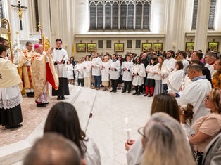 A priest blesses a congregation in white robes holding candles. The ceremony takes place in a bright church with tall windows and golden decor.