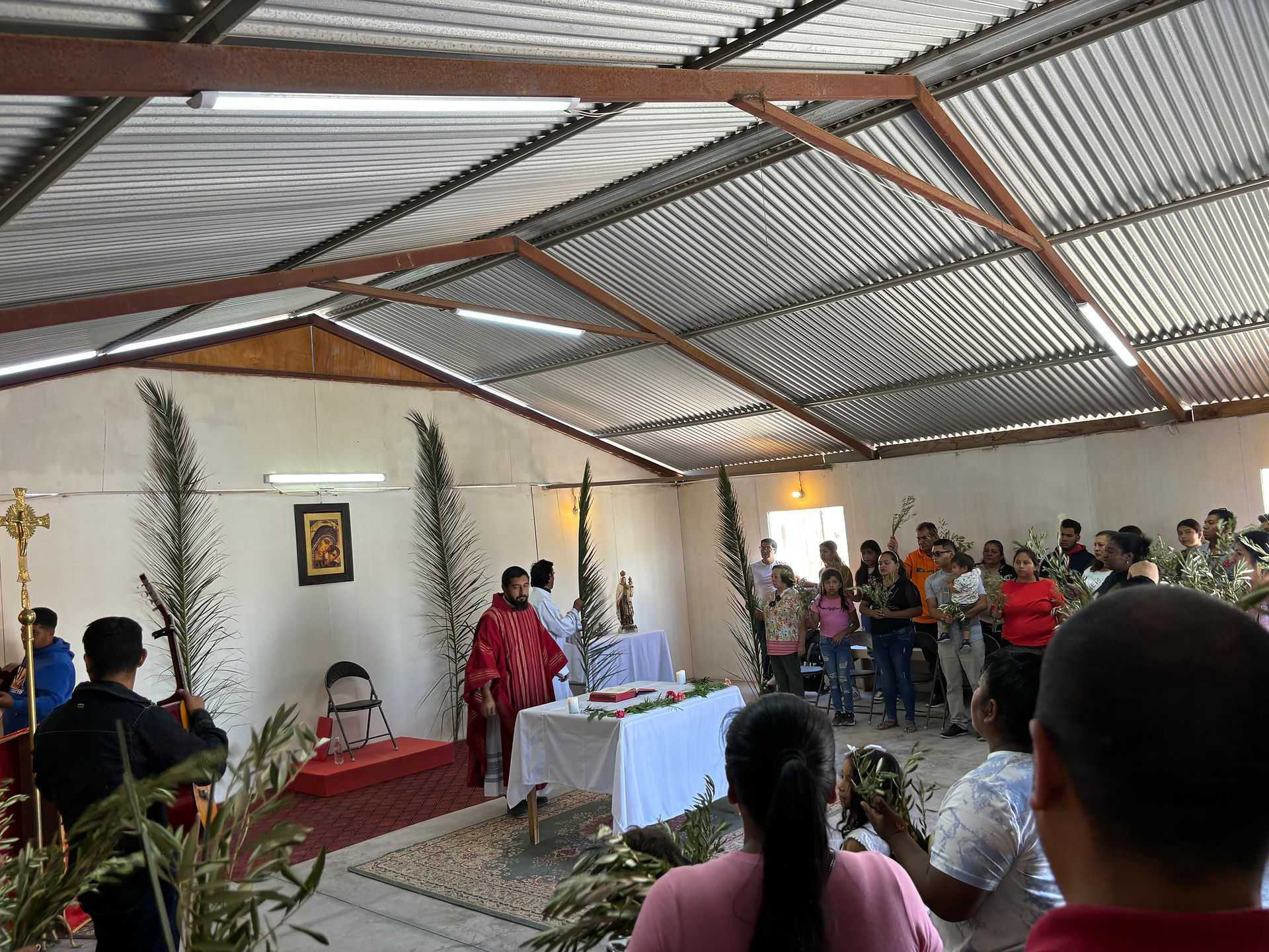People gather in a chapel for a Palm Sunday service. A priest in red stands by a table with a white cloth. Attendees hold palm branches.