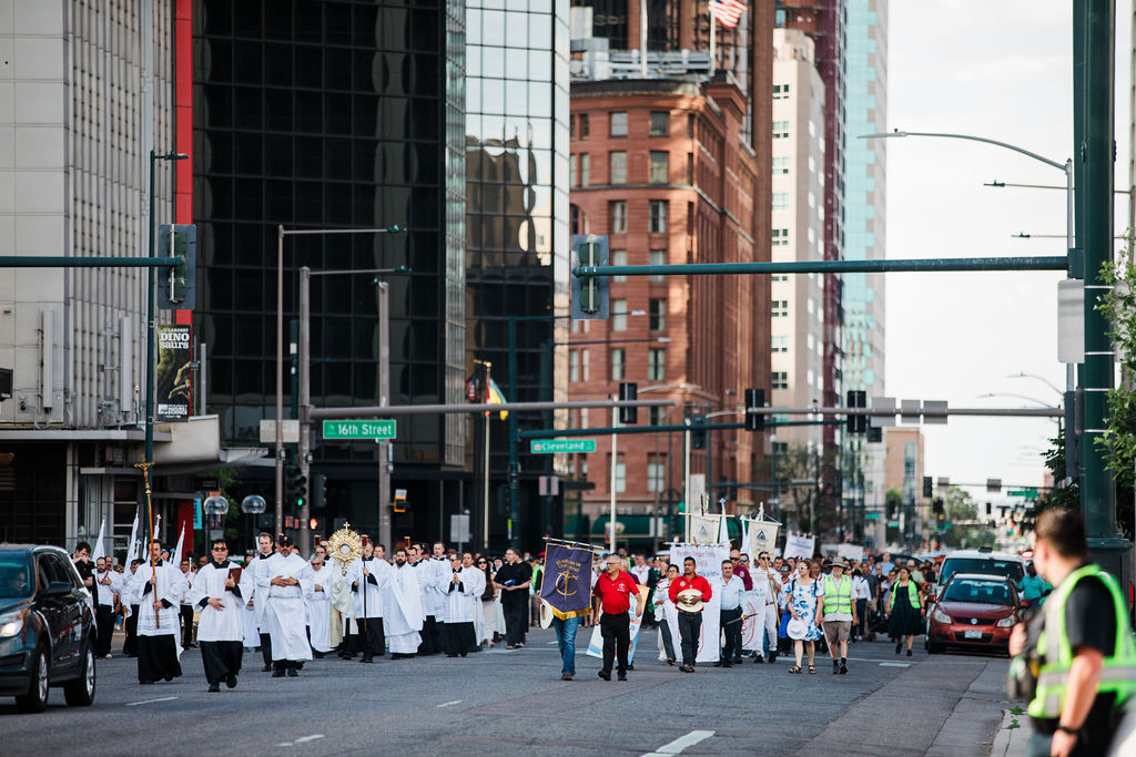 Religious procession of clergy in white robes marches down a downtown street past 16th Street signs and tall buildings.