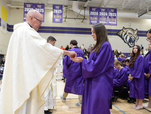 Priest gives communion to a student in purple graduation gown during a ceremony in a gym with banners and a tiger mural.