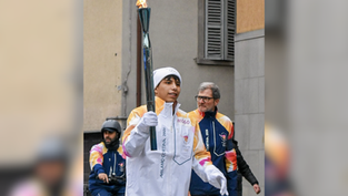 Person carrying a flaming torch, wearing "Milano Cortina 2026" jacket. Another person in similar attire nearby. Urban background, focused mood.