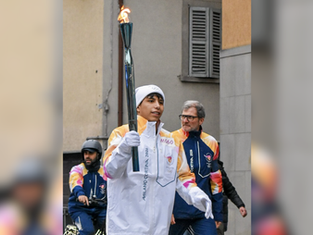 Person carrying a flaming torch, wearing "Milano Cortina 2026" jacket. Another person in similar attire nearby. Urban background, focused mood.