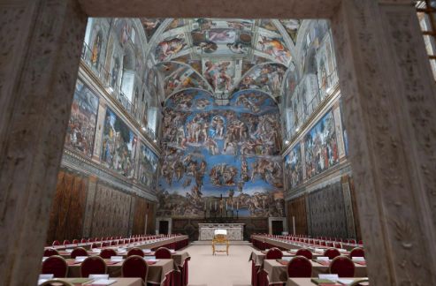 Ornate room with vaulted ceiling, colorful frescoes, and empty chairs lining long tables. Central focus on a prominent chair and altar.