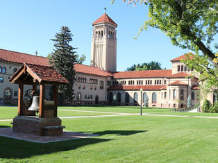 Historic building with red-tiled roofs and bell tower, set on a lush green lawn. A small pavilion with a bell is in the foreground.