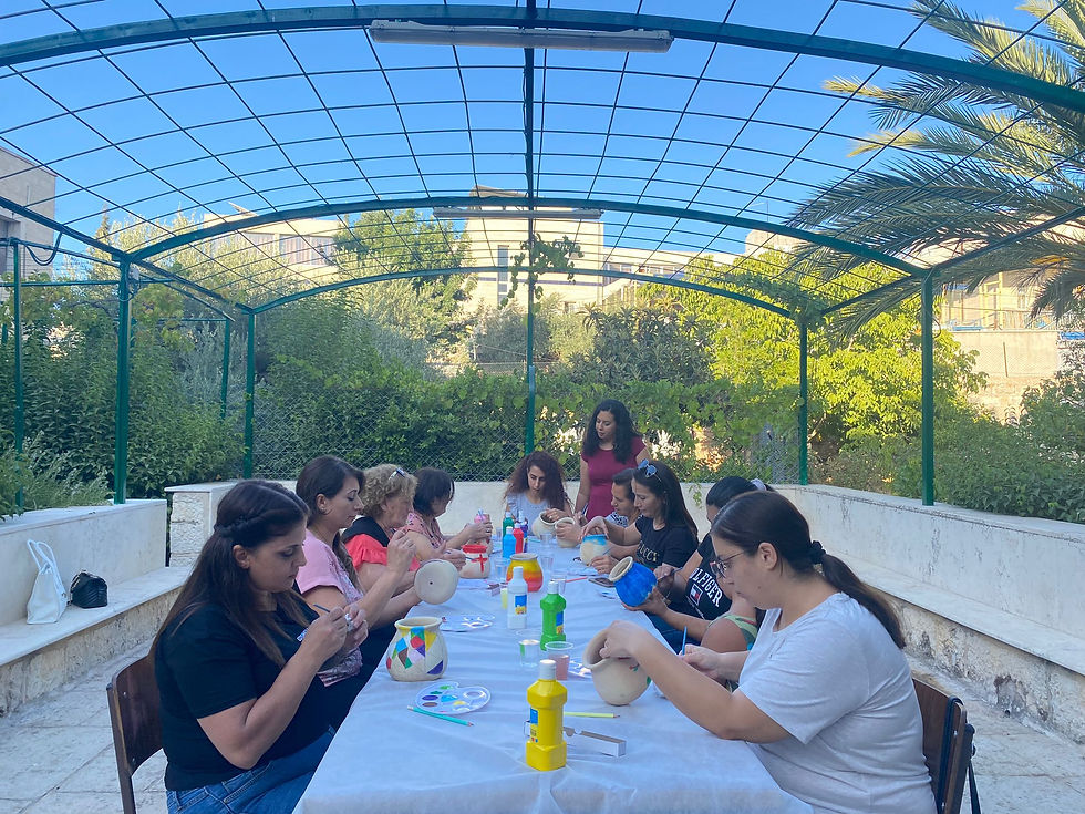 A group of people paint gourds at a long table outdoors under a green pergola. The setting is sunny with green foliage in the background.