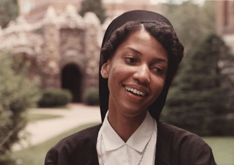 Smiling young nun in black habit and white collar outdoors, with a blurred stone building and garden behind her.