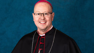 Smiling man in a black clerical robe with red accents, wearing a pink zucchetto. Blue textured background.