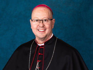 Smiling man in a black clerical robe with red accents, wearing a pink zucchetto. Blue textured background.