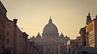 St. Peter's Basilica domes at sunset, framed by buildings. A street with lamps and vehicles leads to the basilica, creating a serene ambiance.
