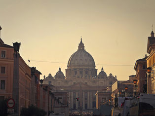 St. Peter's Basilica domes at sunset, framed by buildings. A street with lamps and vehicles leads to the basilica, creating a serene ambiance.