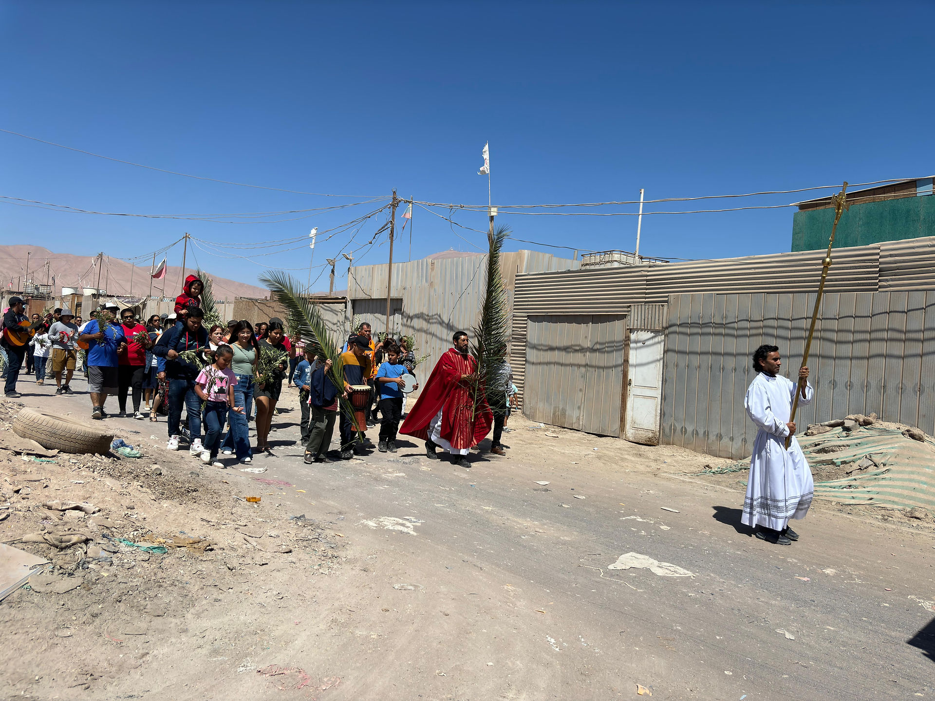People in a procession on a dusty street hold palm leaves. One man in a red robe and another in white lead. Clear blue sky, desert area.