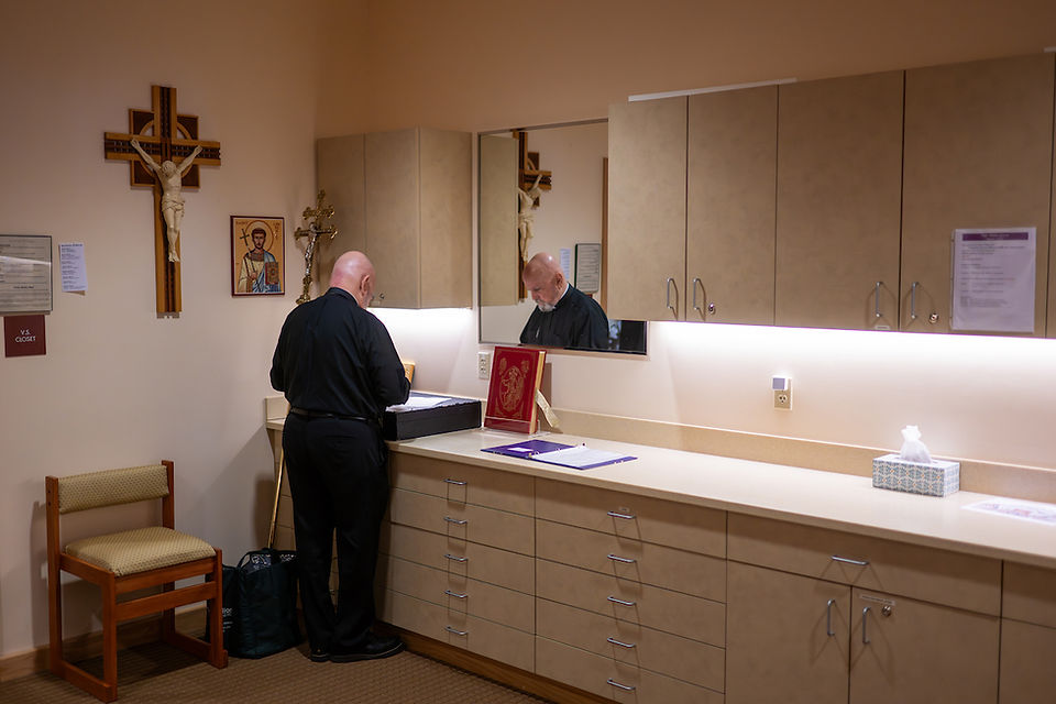 A man in black attire stands in a room with religious icons, looking at a box on a counter. Beige cabinets and a mirror are in the background.