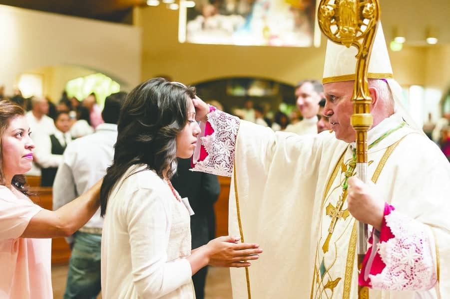 Bishop in white and gold vestments blesses a young woman at a church ceremony, with a blurred crowd in the background.