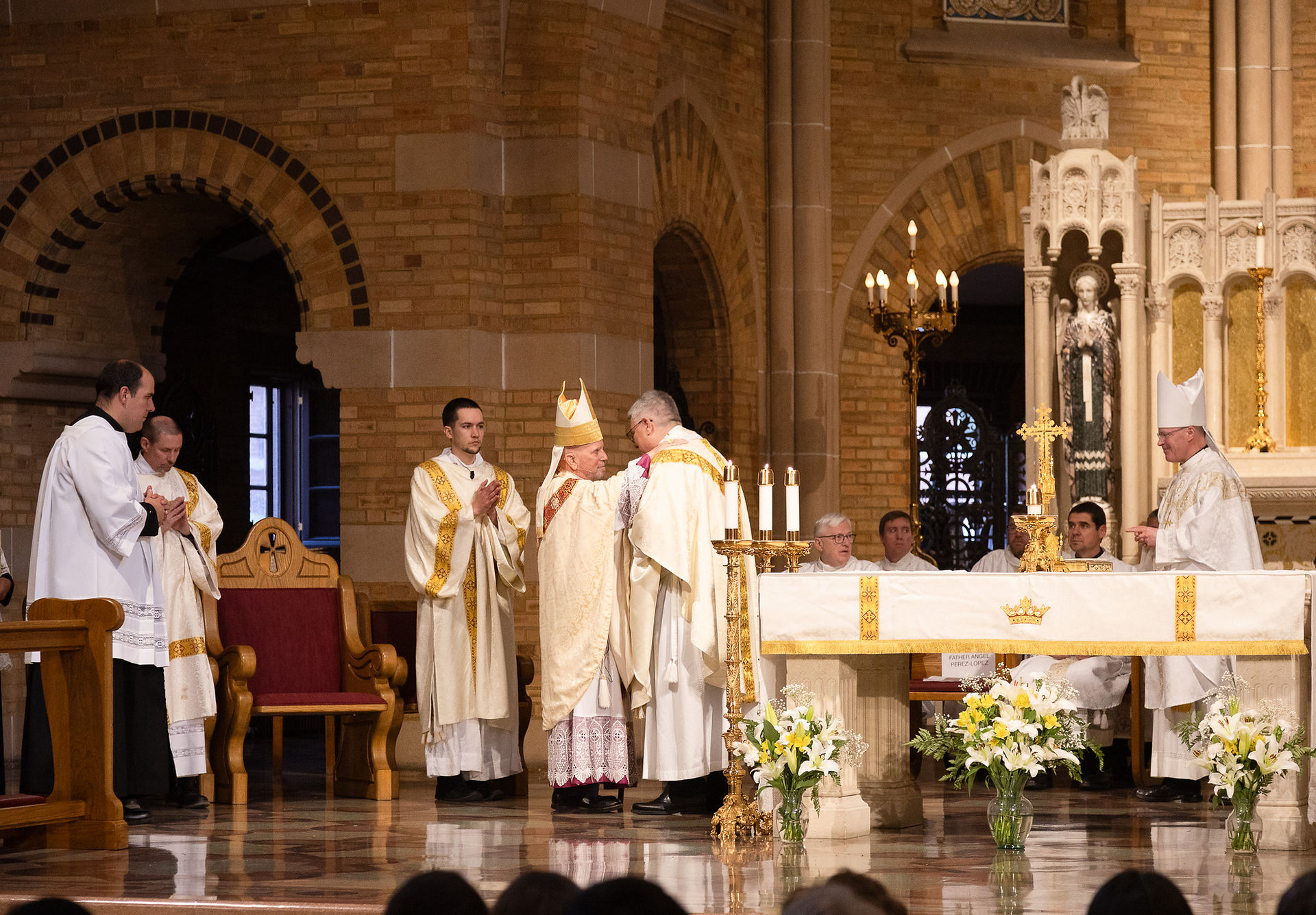 An archbishop embraces a priest during a religious ceremony in an ornate chapel. The mood is celebratory and excited.