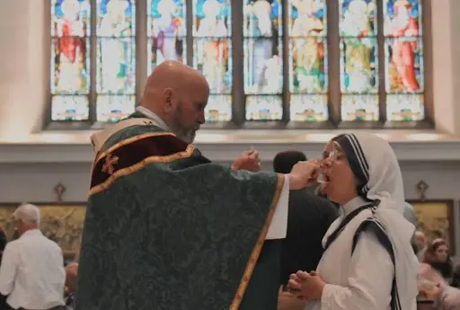 Priest in green robe gives communion to a nun in white, inside a church with colorful stained glass windows. Spiritual and solemn mood.