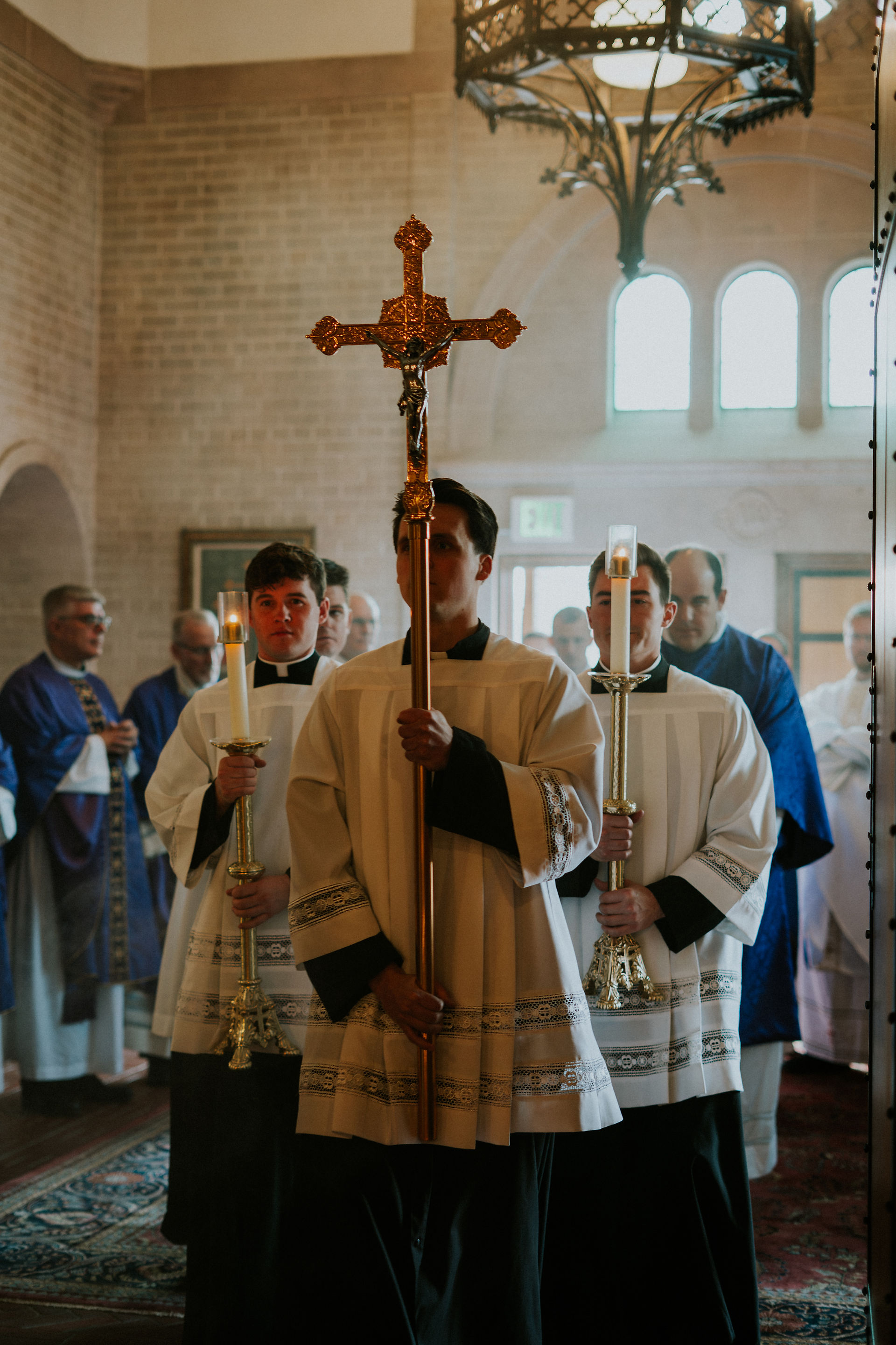 Clerics in white robes solemnly hold candles and a cross, processing in a church with arched windows. Others in purple robes follow.