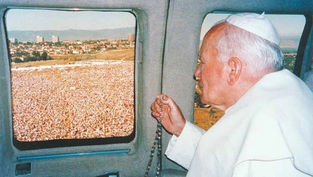 Religious leader in white robes holding a rosary while looking out a helicopter window at a large outdoor crowd below.