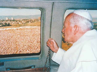 Religious leader in white robes holding a rosary while looking out a helicopter window at a large outdoor crowd below.