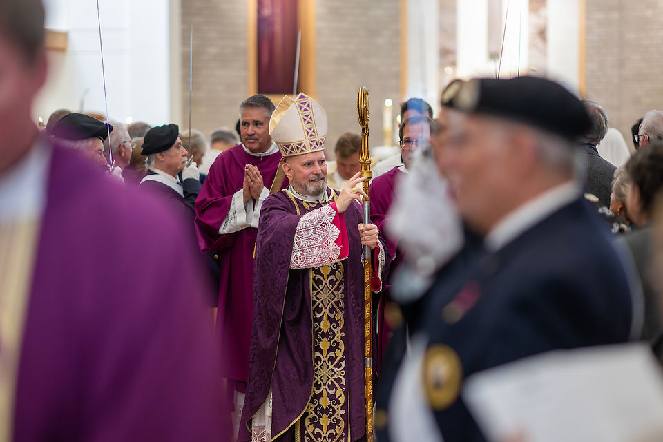 A bishop in ornate purple robes holds a staff, leading a procession in a church setting. Clergy in similar attire follow, displaying solemnity.