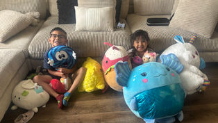 Two kids sit on a beige couch with large plush toys, smiling. The wooden floor contrasts with bright toy colors, creating a joyful mood.