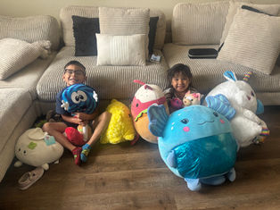 Two kids sit on a beige couch with large plush toys, smiling. The wooden floor contrasts with bright toy colors, creating a joyful mood.