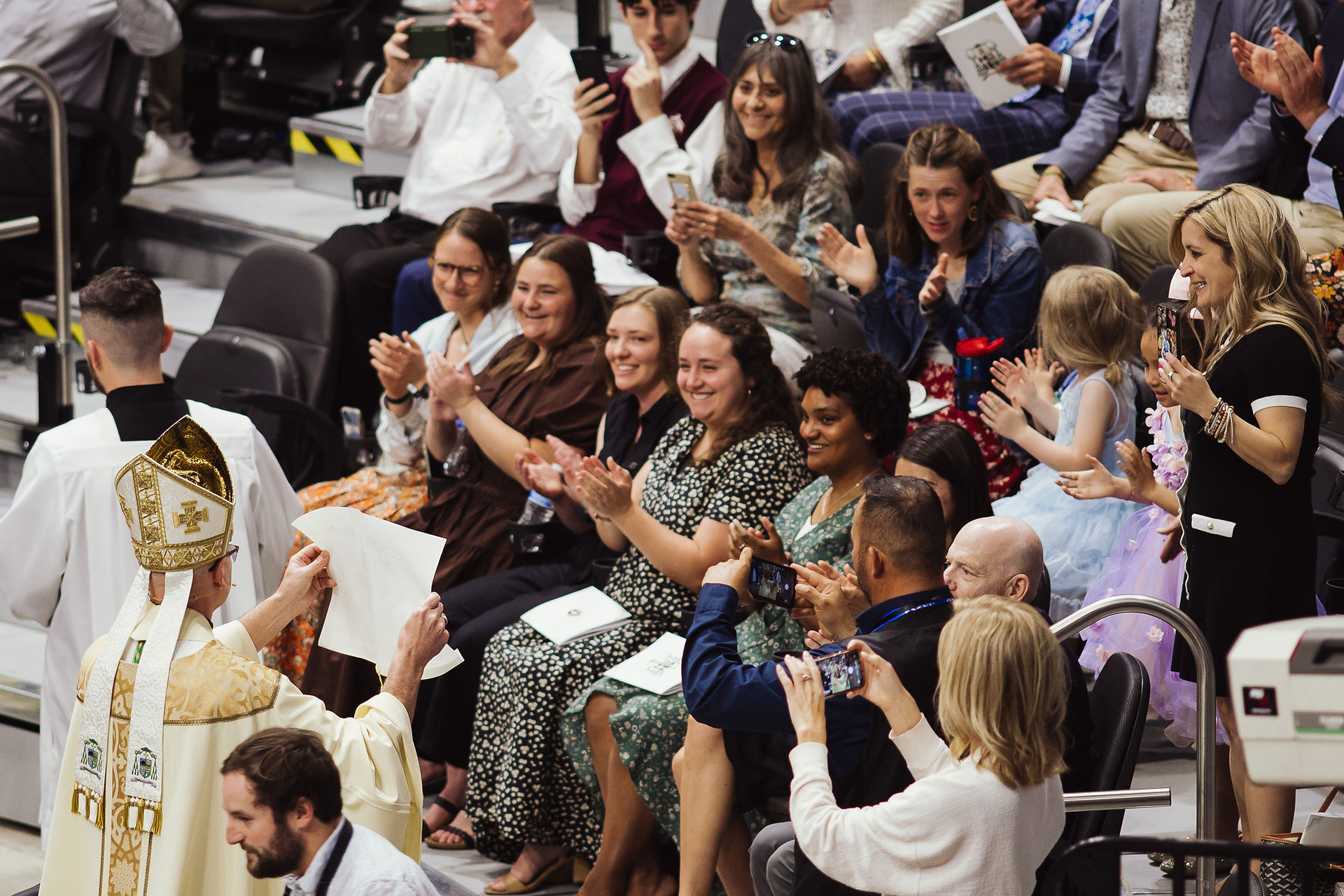 A bishop in ceremonial attire presents a document to a seated audience. People clap and take photos in an indoor setting, conveying joy.