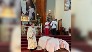 A priest conducts a funeral service in a church, standing beside a casket. Altar servers stand behind. Christmas trees and religious art decorate the background.