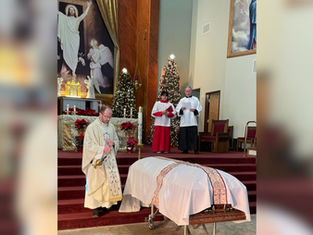 A priest conducts a funeral service in a church, standing beside a casket. Altar servers stand behind. Christmas trees and religious art decorate the background.