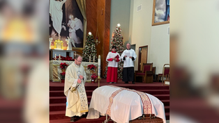A priest conducts a funeral service in a church, standing beside a casket. Altar servers stand behind. Christmas trees and religious art decorate the background.