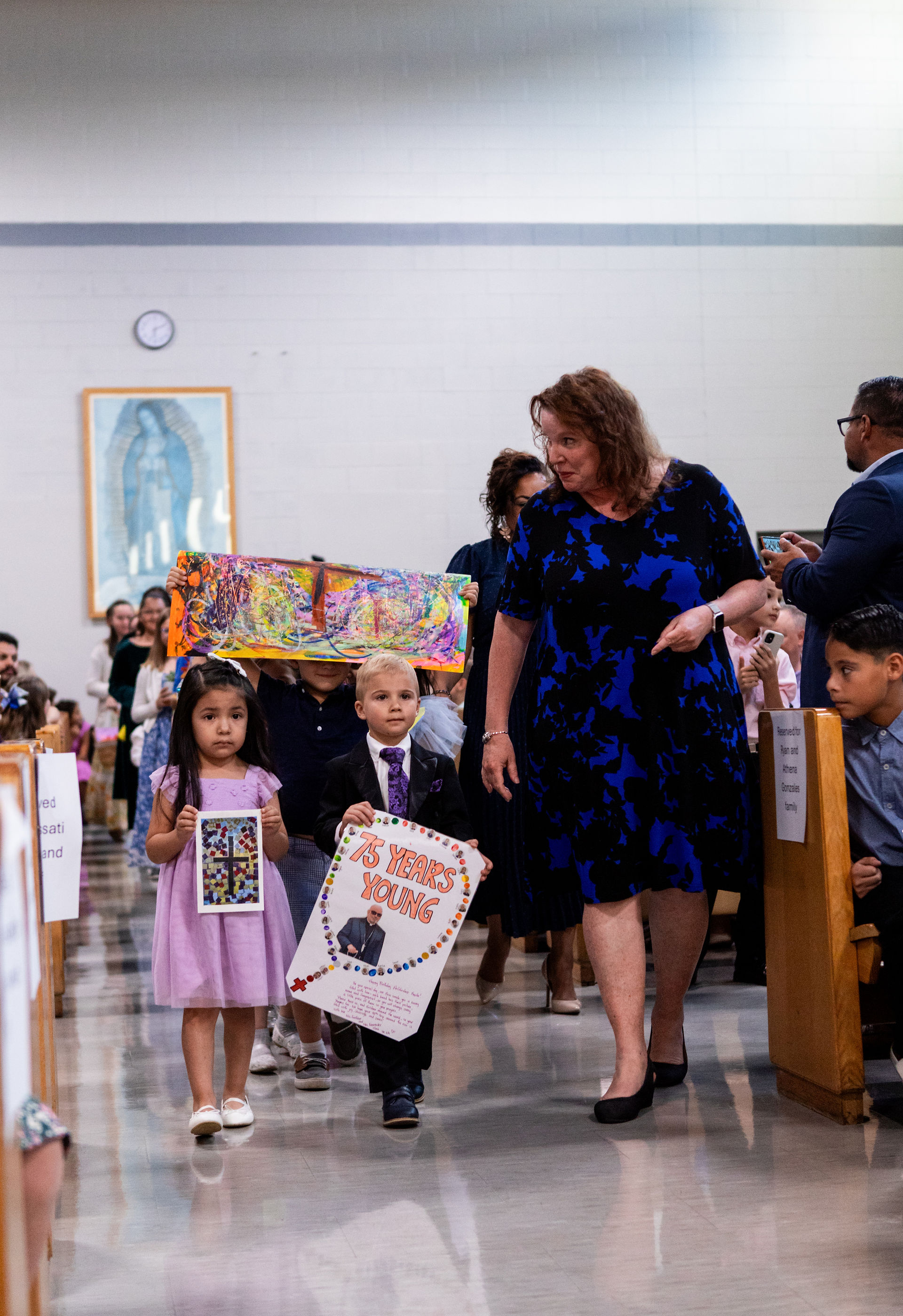Children and a woman in a church aisle holding colorful signs, one reads "75 Years Young." A crowd watches, creating a festive atmosphere.